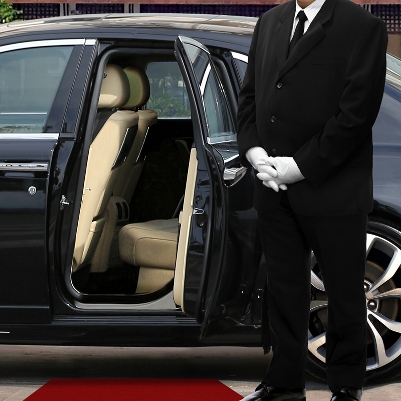 Chauffeur en costume noir et gants blancs ouvre la portière d'une voiture de luxe noire avec intérieur beige sur un tapis rouge.