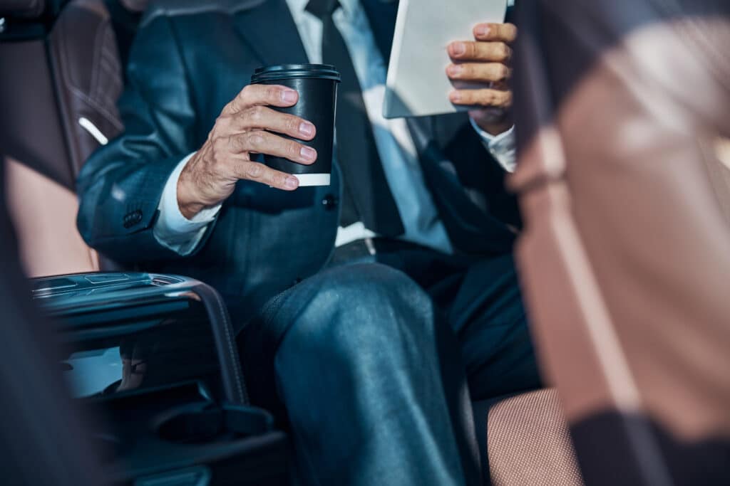 Homme d'affaires en costume sombre tenant une tasse de café noir et une tablette blanche, assis dans une voiture de luxe aux sièges en cuir.