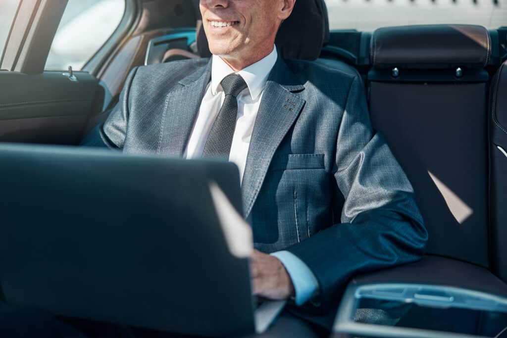 Homme d'affaires souriant en costume utilise un ordinateur portable à l'arrière d'une voiture de luxe, en pleine lumière du jour.
