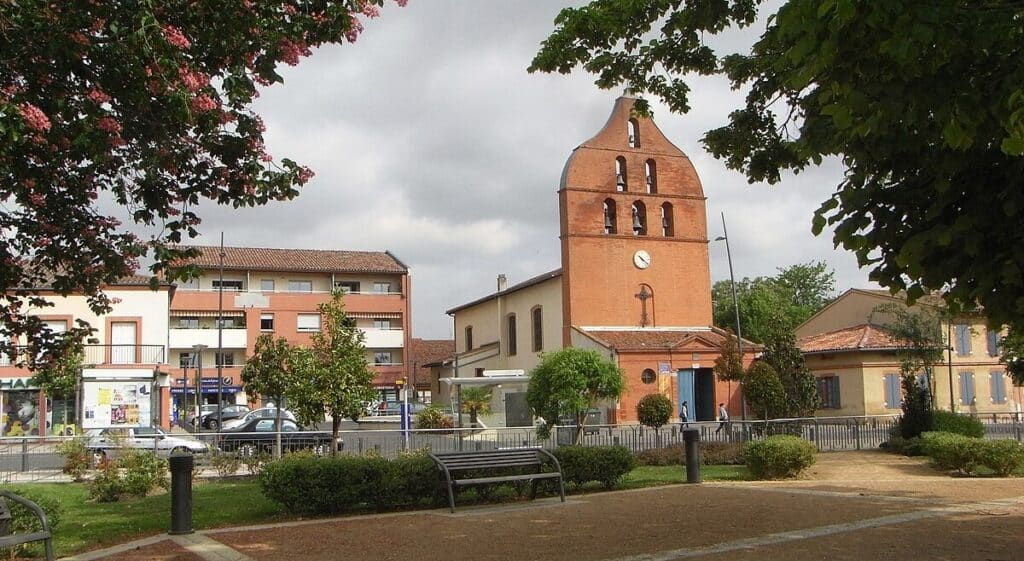 Église au clocher de briques rouges dominant une place de ville verdoyante avec bancs, bâtiments, commerces dont une Pharmacie, et voitures.